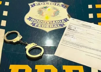 A imagem mostra o capô de uma viatura da Polícia Rodoviária Federal (PRF) e, em cima dele, um mandado de prisão. (Foto: Divulgação | PRF)