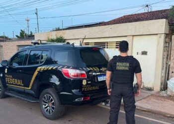 A imagem em destaque mostra um policial federal próximo a uma viatura da instituição e a um dos locais alvos dos mandados. (Foto: Divulgação | Ascom PF)